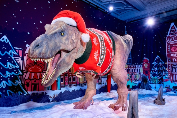 A large animatronic T-rex wearing a festive jumper and Santa hat. The background is an illustration of snow covered buildings